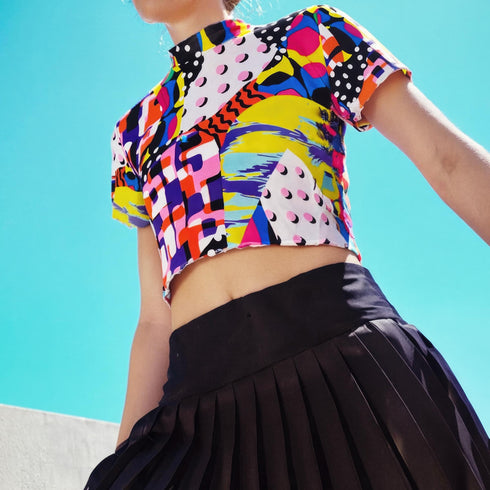 A colorful 90s-style mesh crop top featuring a maximalist abstract pattern. This see-through shirt has a high mock neck, short sleeves, and a vibrant, retro design with geometric shapes, color blocking, and polka dots in bold shades of pink, yellow, blue, and orange. The Y2K-inspired top is a statement piece of festival clothing, perfect for a rave outfit or alternative, dopamine-dressing-inspired streetwear looks.